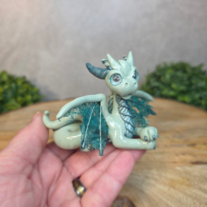A hand holding an aqua and turquoise clay dragon against a blurred wood surface
