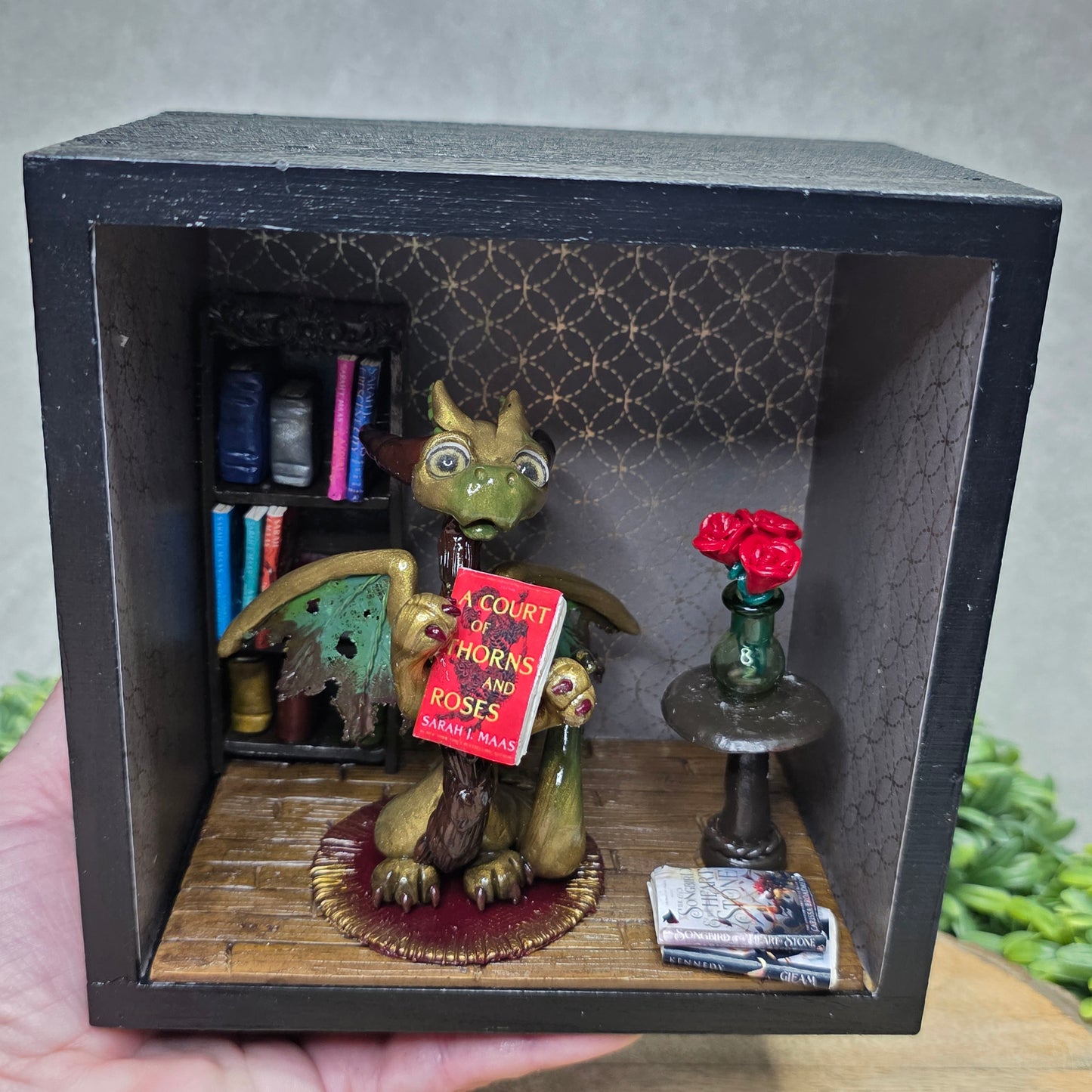 Hand holding a Book nook with dragon on a wood surface with greenery