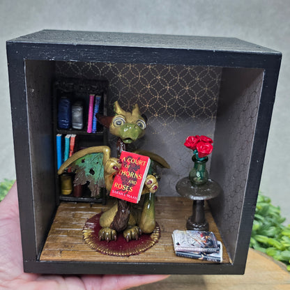 Hand holding a Book nook with dragon on a wood surface with greenery