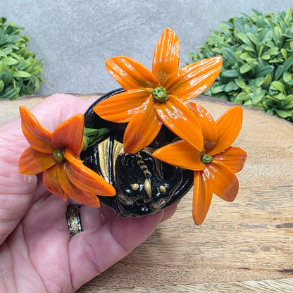 Top view of a hand holding a clay baby dragon in a pot with lilies against a wood surface with greenery.