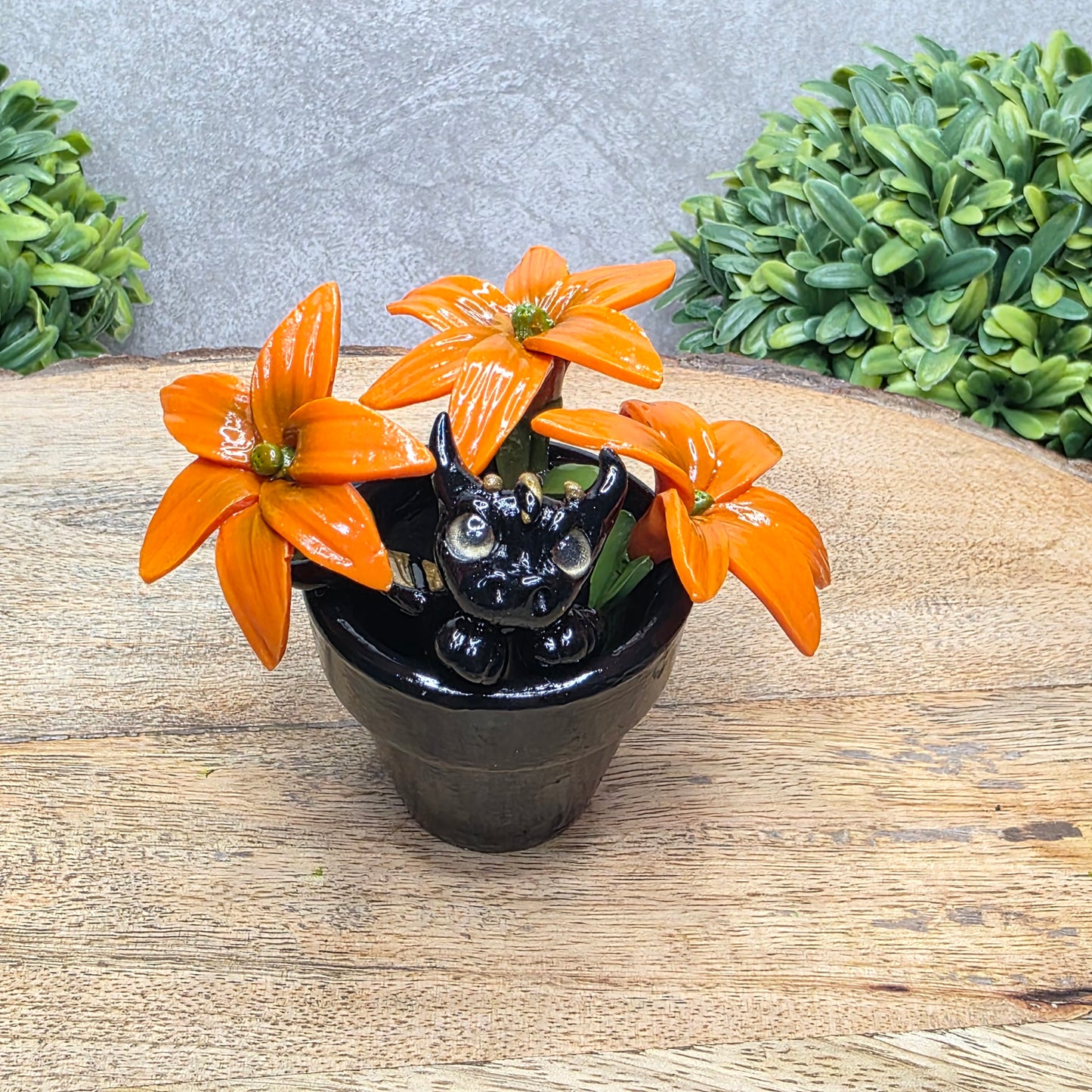 A clay baby dragon in a pot with lilies against a wood surface with greenery.