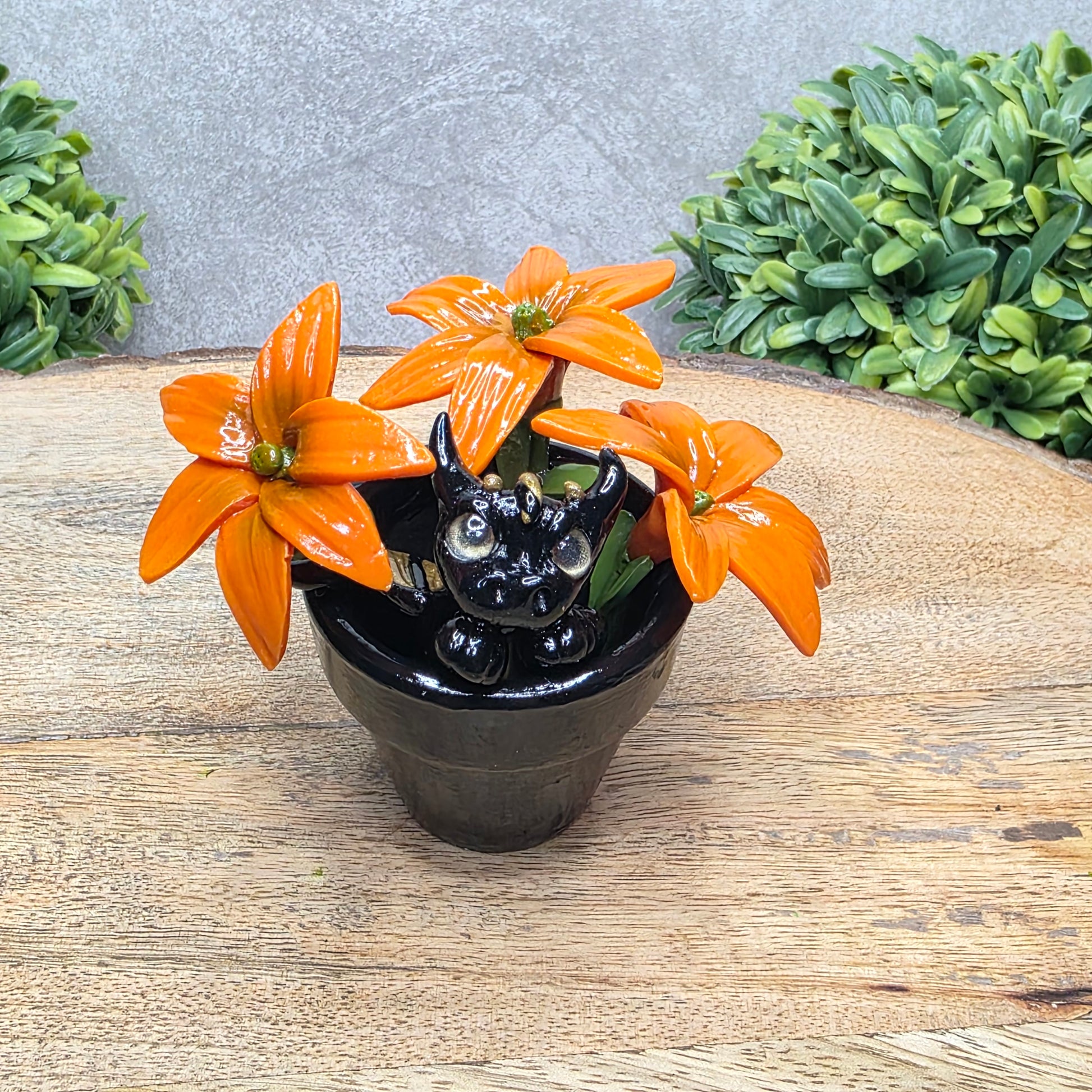 A clay baby dragon in a pot with lilies against a wood surface with greenery.
