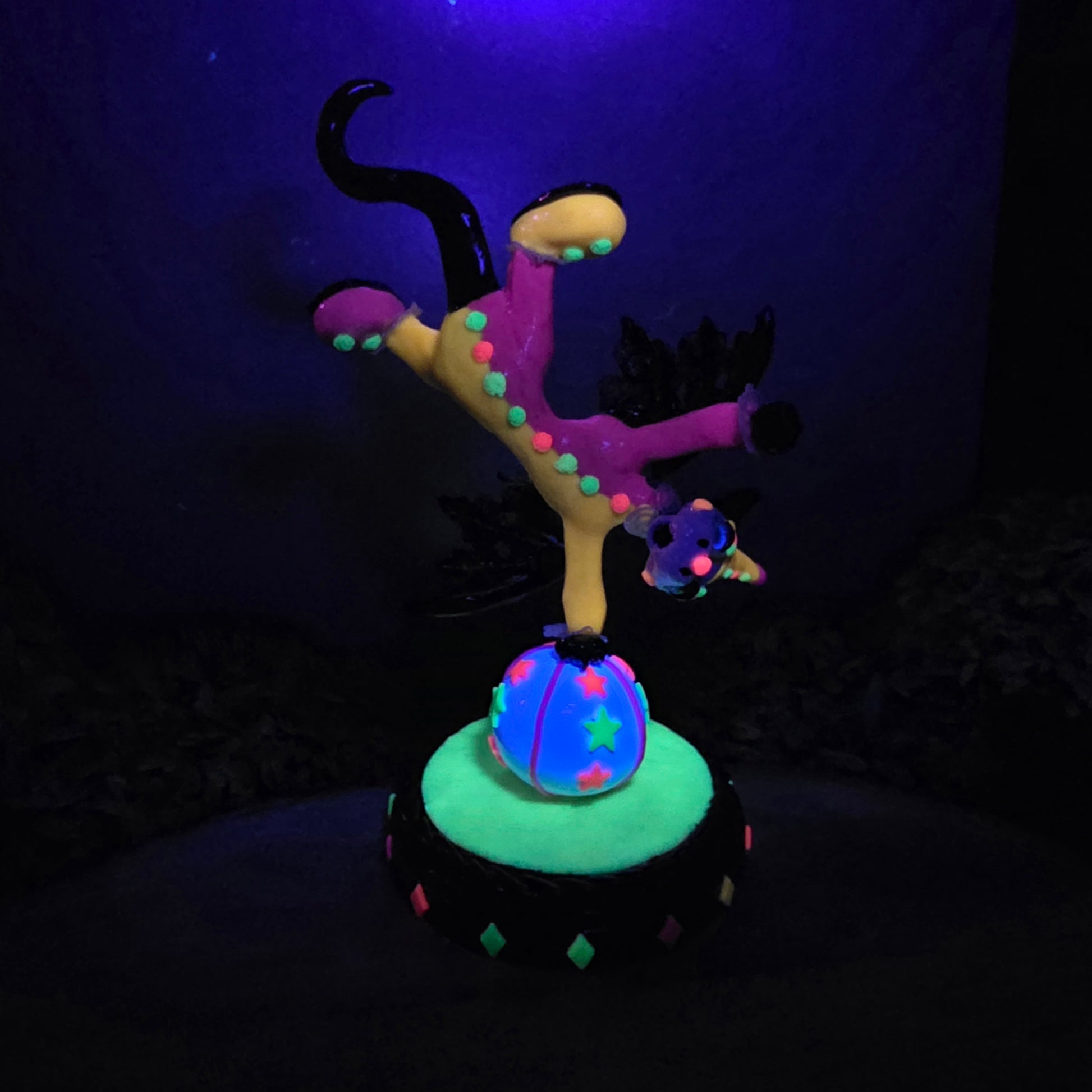 A blacklight reactive clown dragon balancing on a ball on a wood surface with greenery