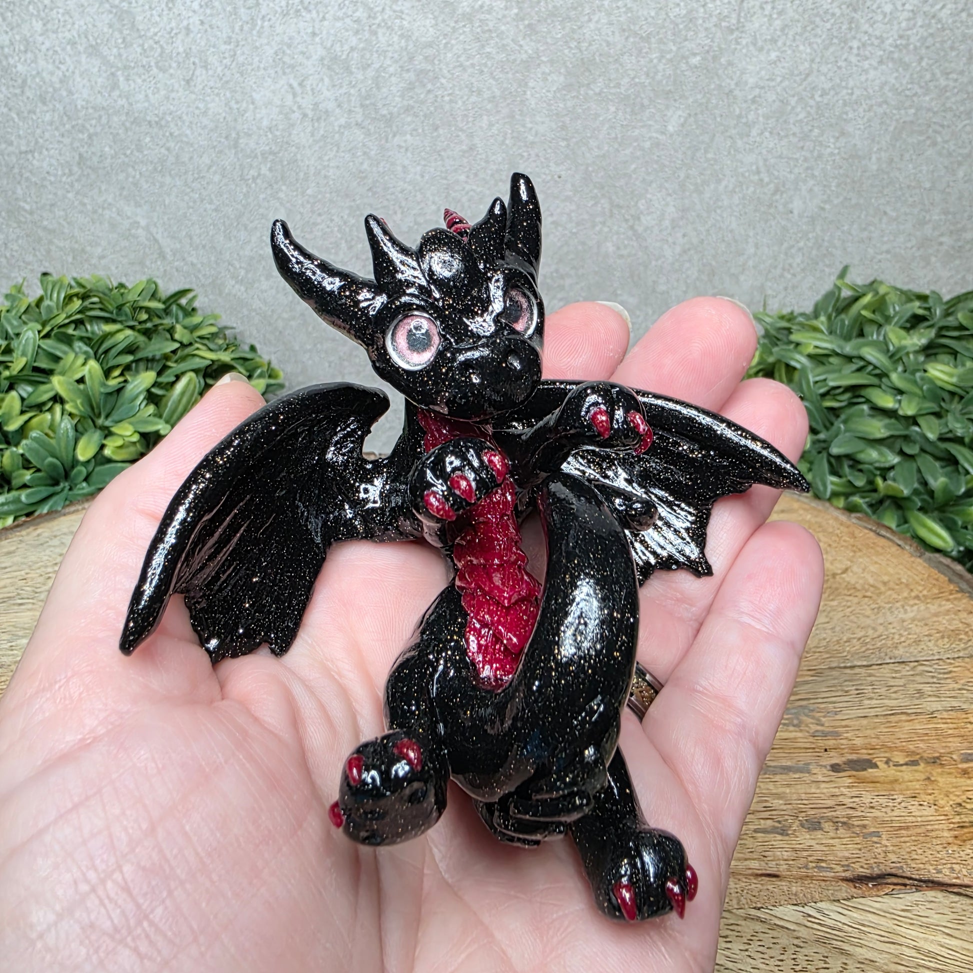A hand holding a clay dragon against a wood background with greenery