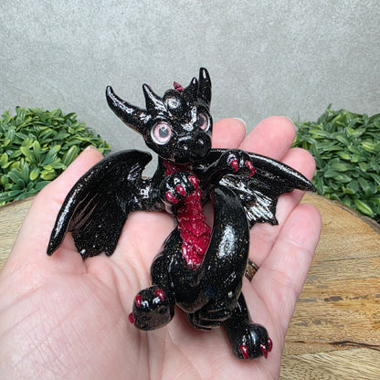 A hand holding a clay dragon against a wood background with greenery