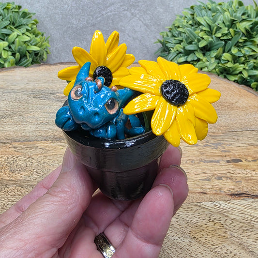 Hand holding a clay baby dragon in a pot with black eye susans against a wood surface with greenery.