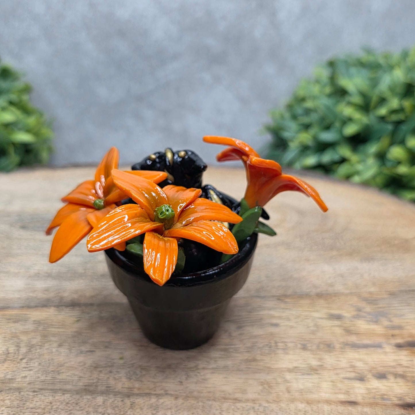 Back view of a clay baby dragon in a pot with lilies against a wood surface with greenery.