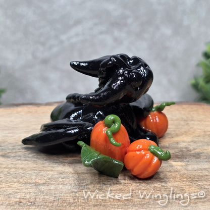 A baby clay dragon sculpture with pumpkins against a wood background with greenery and text Wicked Winglings