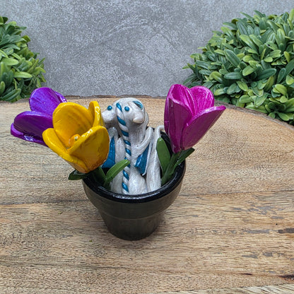 Back view of a clay baby dragon in a pot with tulips against a wood surface with greenery.