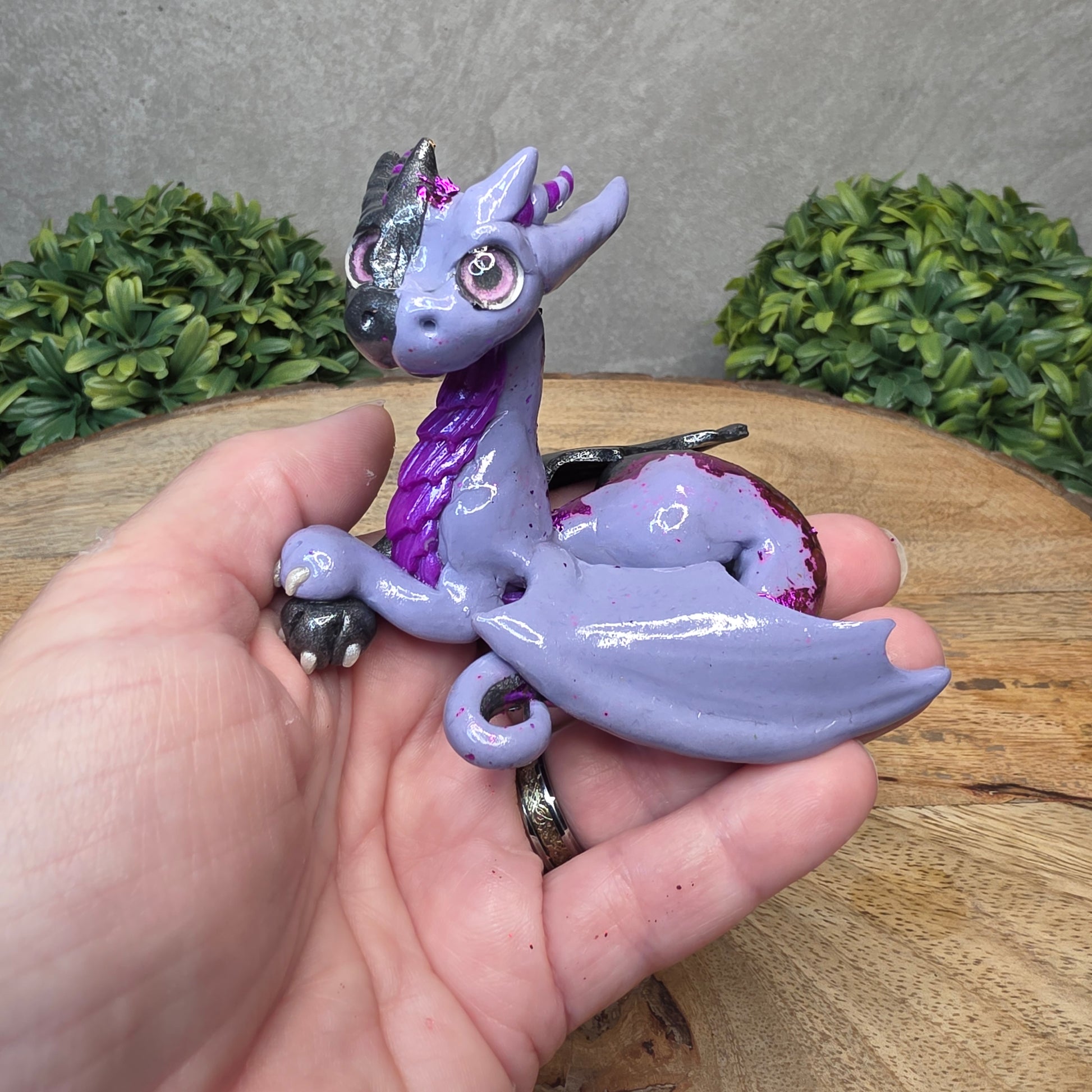 A hand holding a 2 toned clay dragon sculpture on a wood surface with greenery.