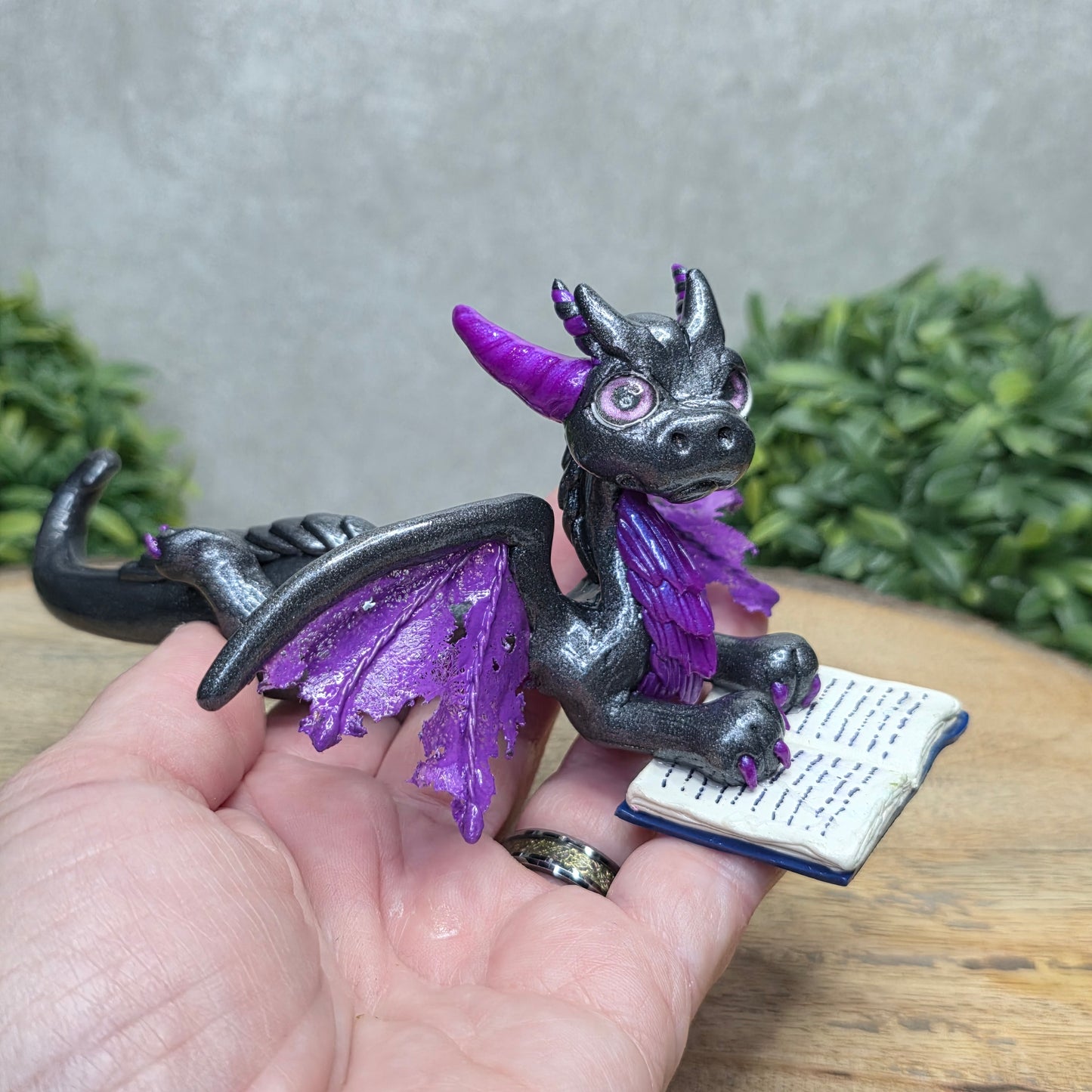 Hand holding a dragon reading a book against a wood surface with greenery