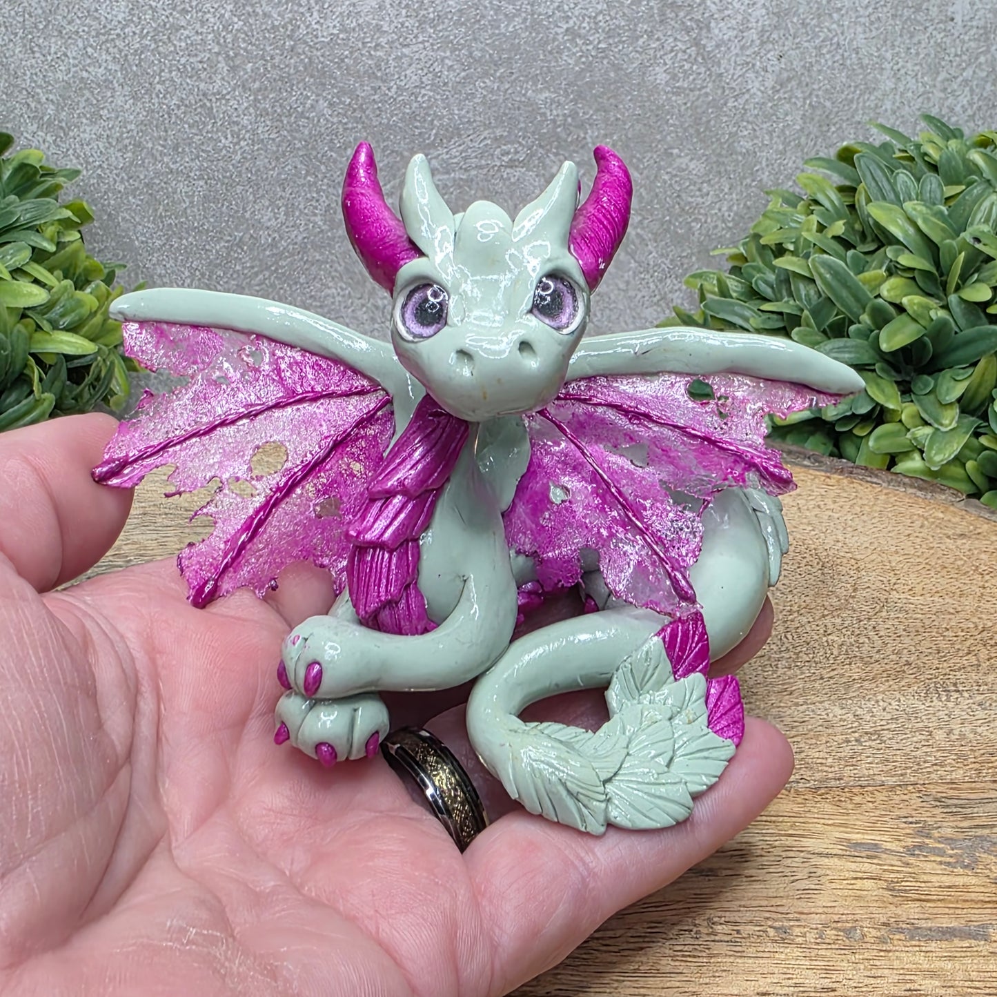 Hand holding a clay dragon against a wood surface with greenery.