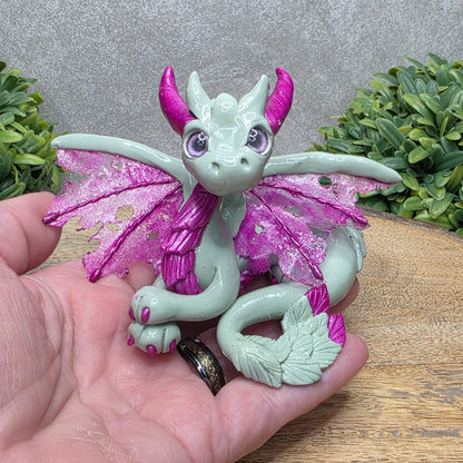Hand holding a clay dragon against a wood surface with greenery.