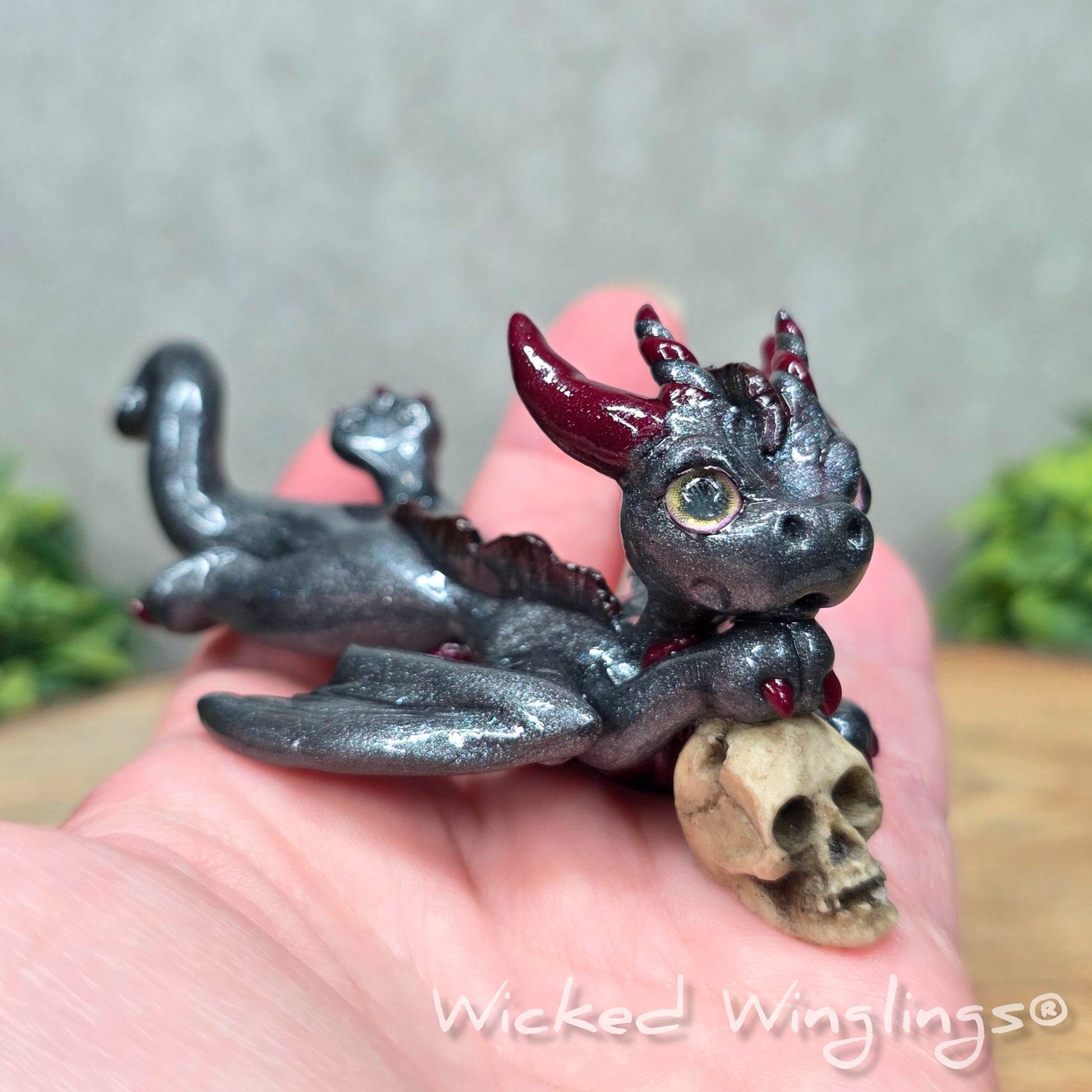 A hand holding a clay dragon with a skull against a wood surface with greenery. Includes text Wicked Winglings