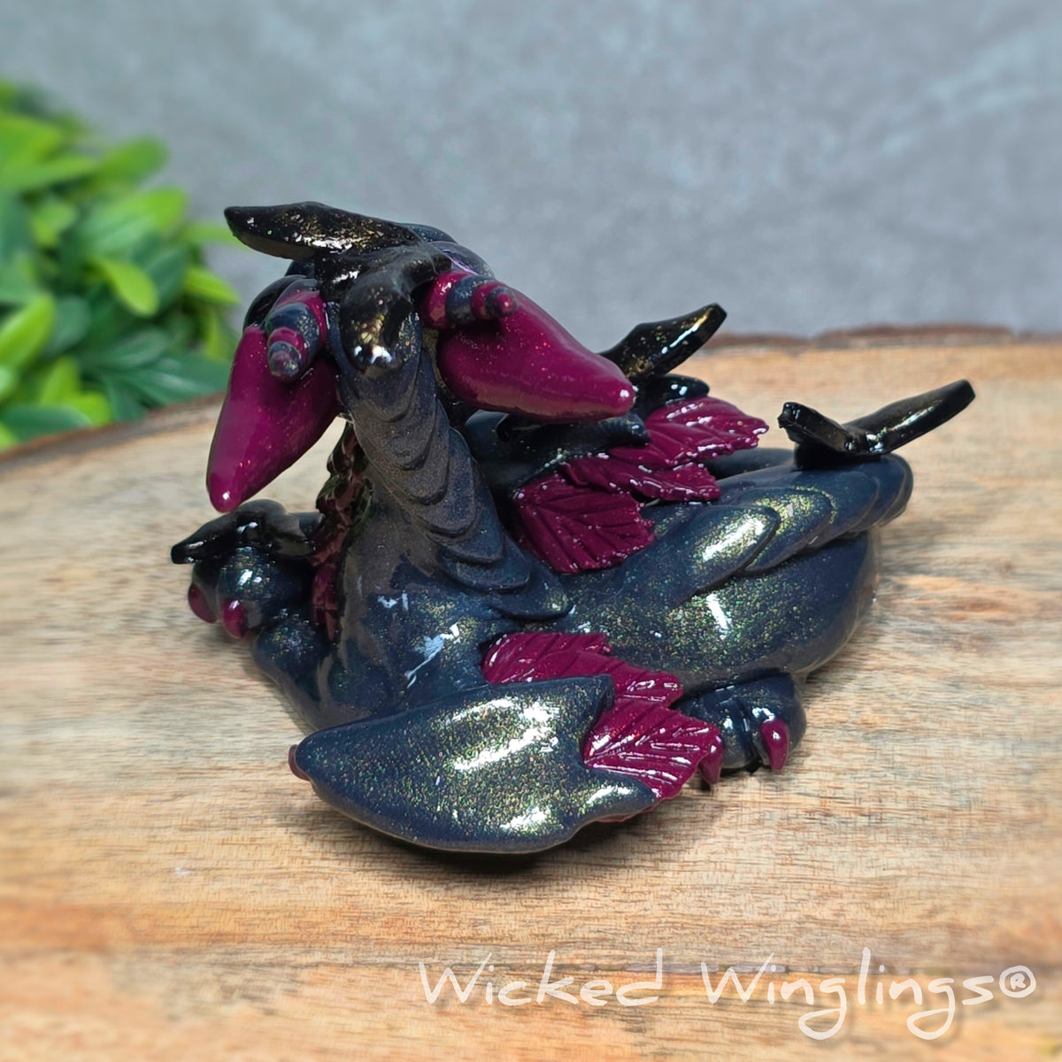 Back view of a clay dragon with bats sitting on a wood surface with greenery. Text reads Wicked Winglings.