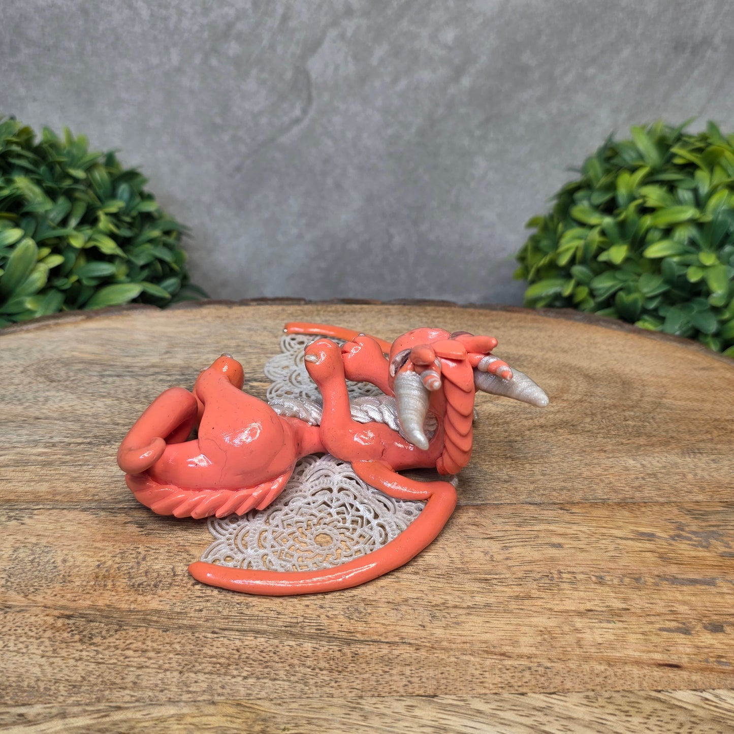 A clay dragon sculpture with lacy wings on a wood surface with greenery.