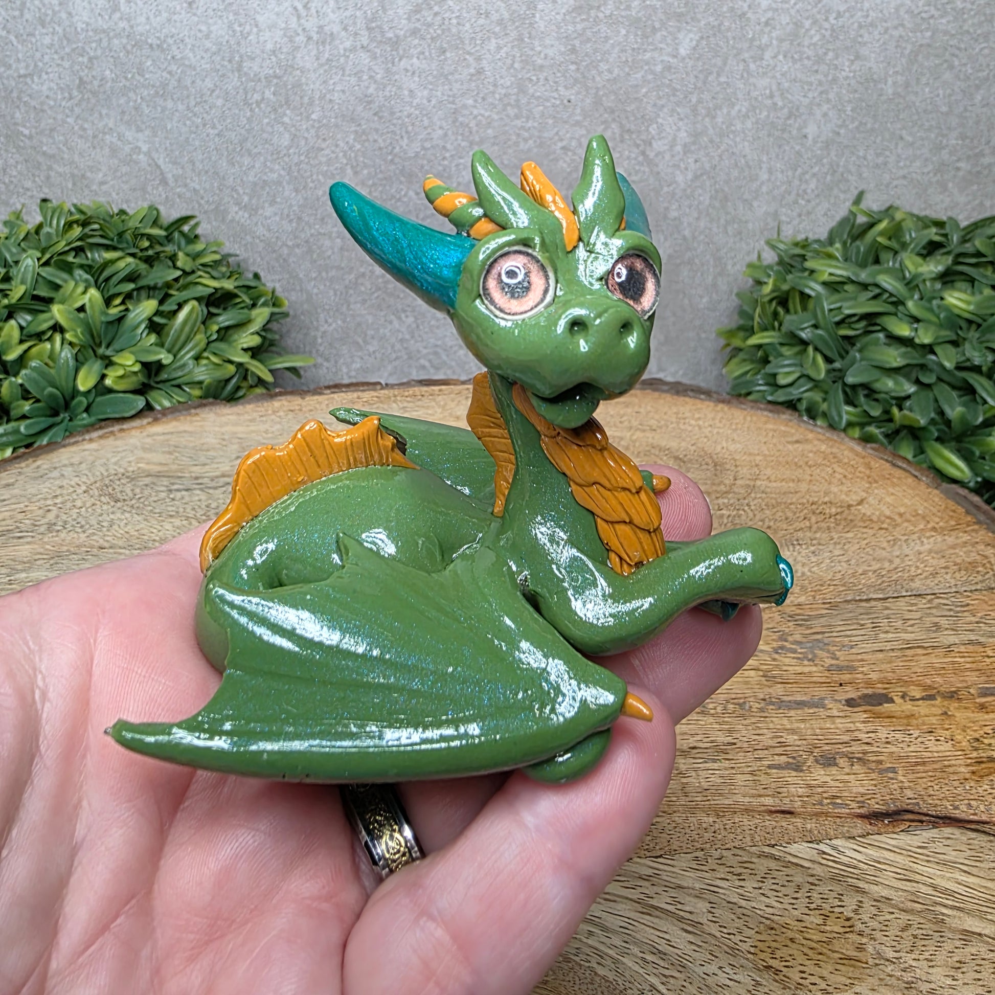 A hand holding a clay dragon against a wood background with greenery