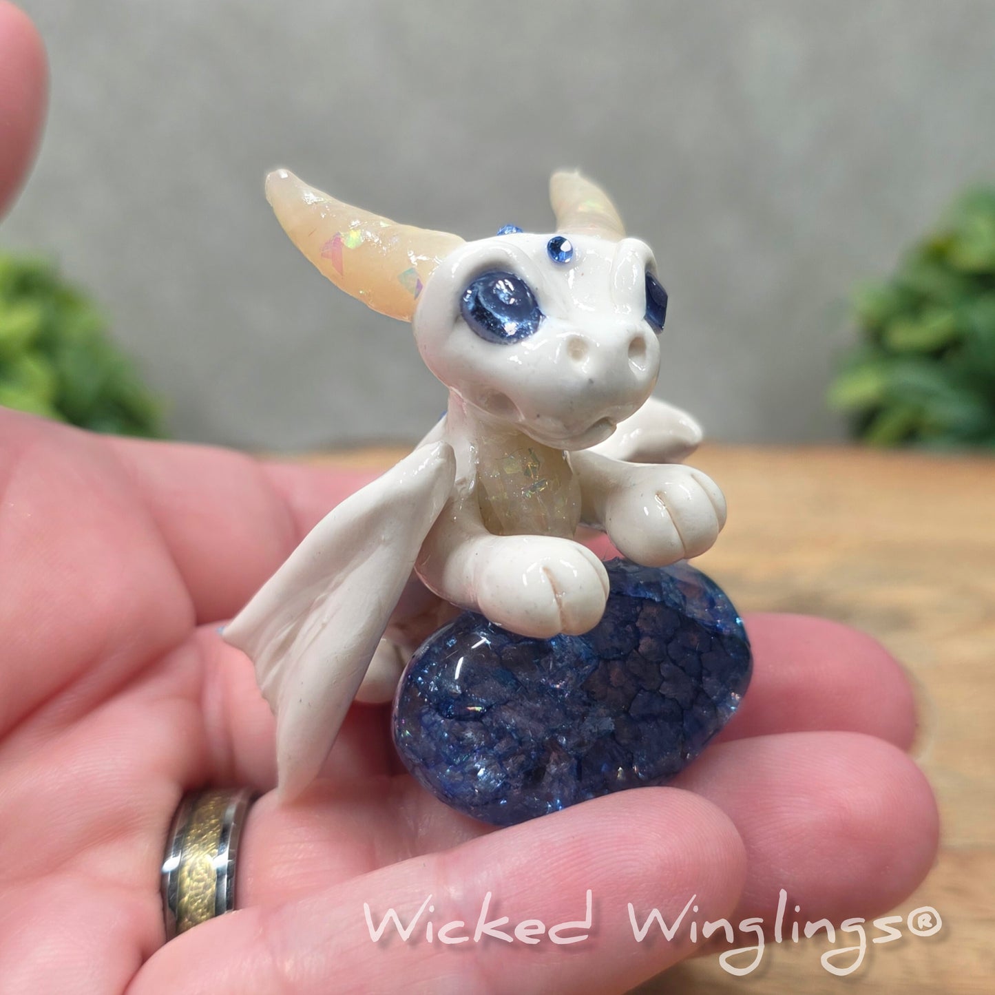 A hand holding a baby clay dragon sculpture with a crackled quartz against a wood background with greenery and text Wicked Winglings