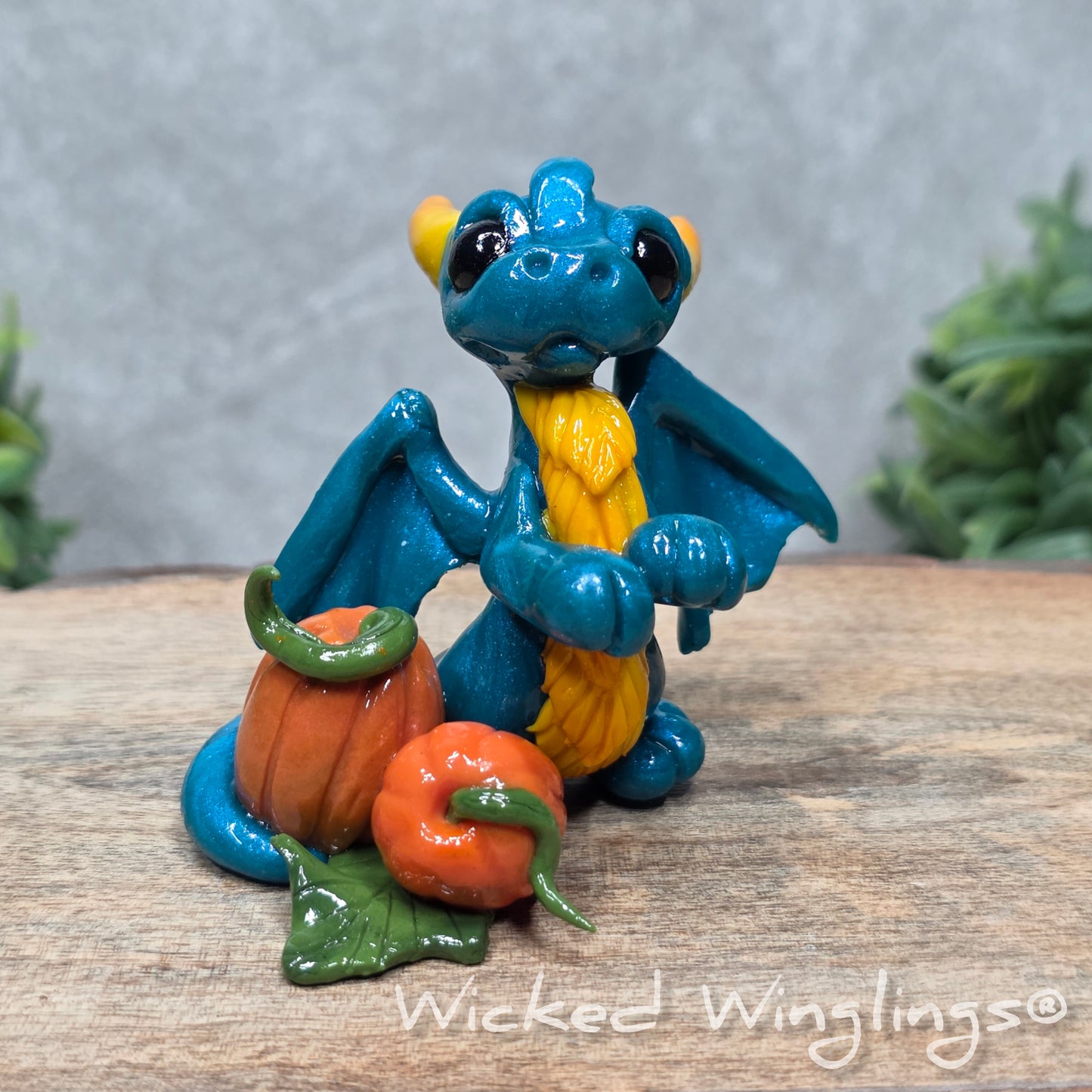 A baby clay dragon sculpture with pumpkins against a wood background with greenery and text Wicked Winglings
