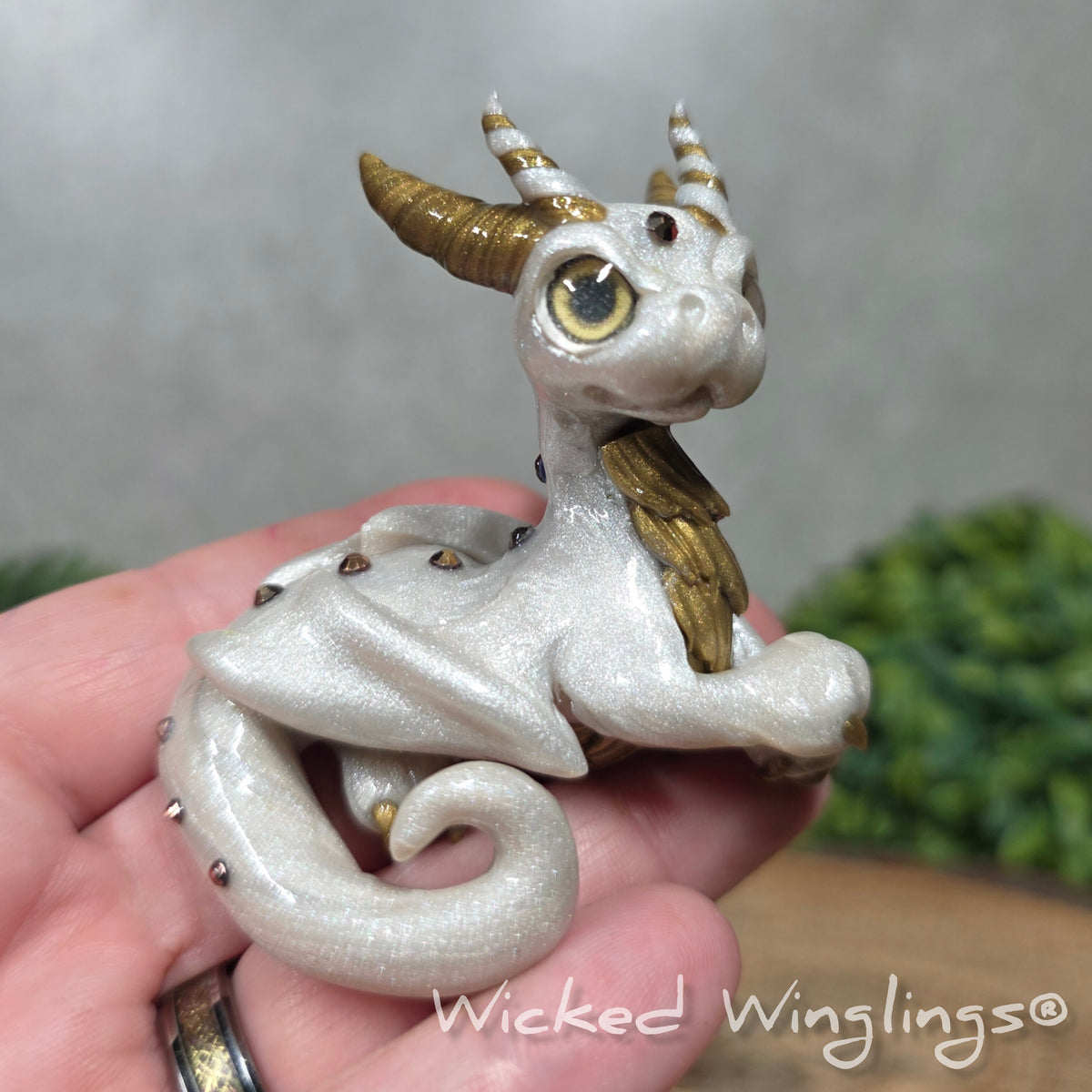 A hand holding a white clay dragon against a blurred background with greenery. Text says Wicked Winglinng.