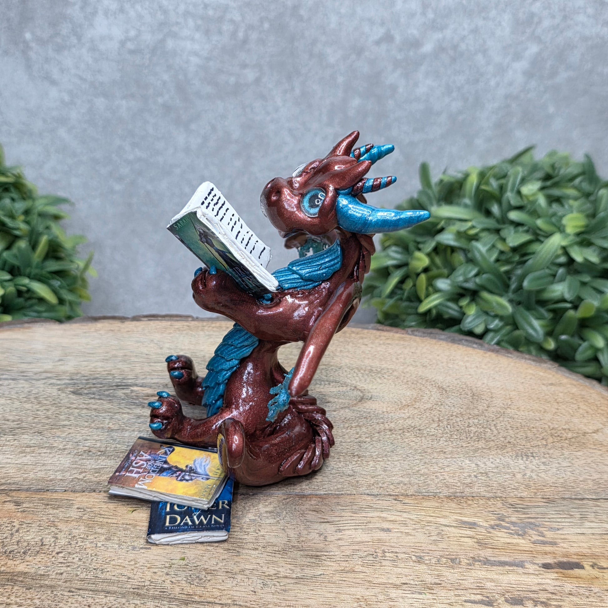 A dragon reading a book against a wood surface with greenery