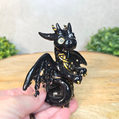 A hand holding a clay dragon figurine on a wooden surface with a blurred green plant in the background