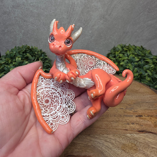 A hand holding a clay dragon sculpture with lacy wings on a wood surface with greenery.