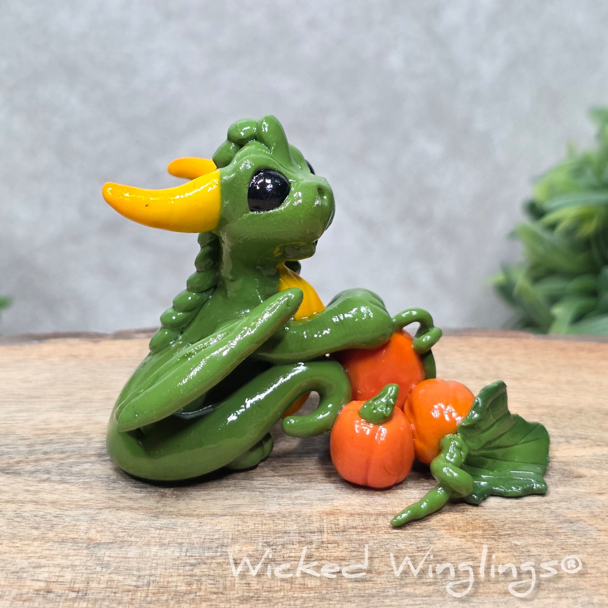 A baby clay dragon sculpture with pumpkins against a wood background with greenery and text Wicked Winglings