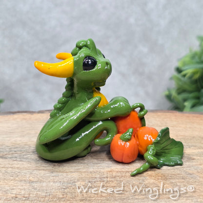 A baby clay dragon sculpture with pumpkins against a wood background with greenery and text Wicked Winglings