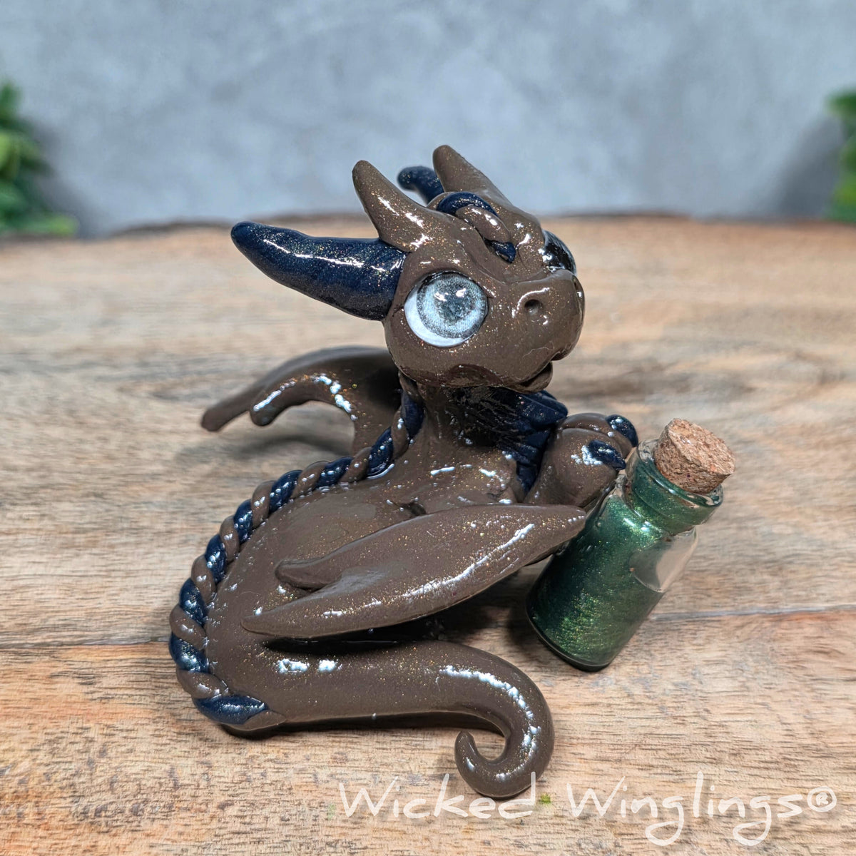 A clay dragon with a potion bottle on a wood surface. Text reads Wicked Winglings.