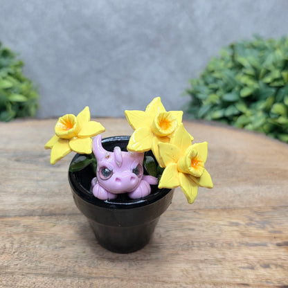 A clay baby dragon in a pot with daffodils against a wood surface with greenery.