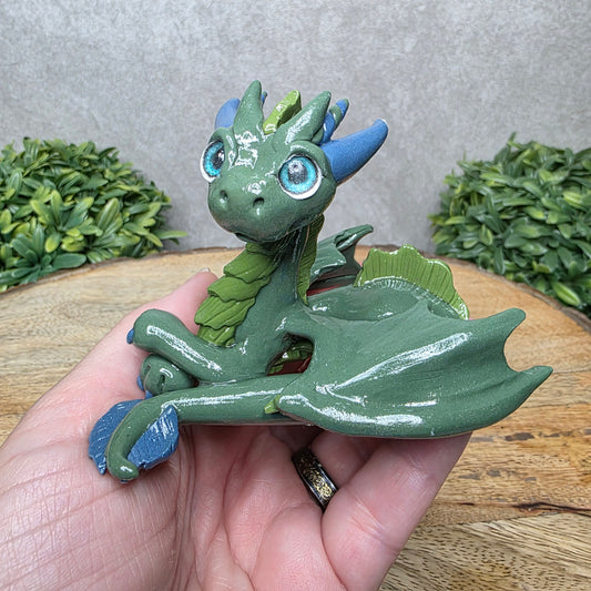 A hand holding a clay dragon against a wood background with greenery