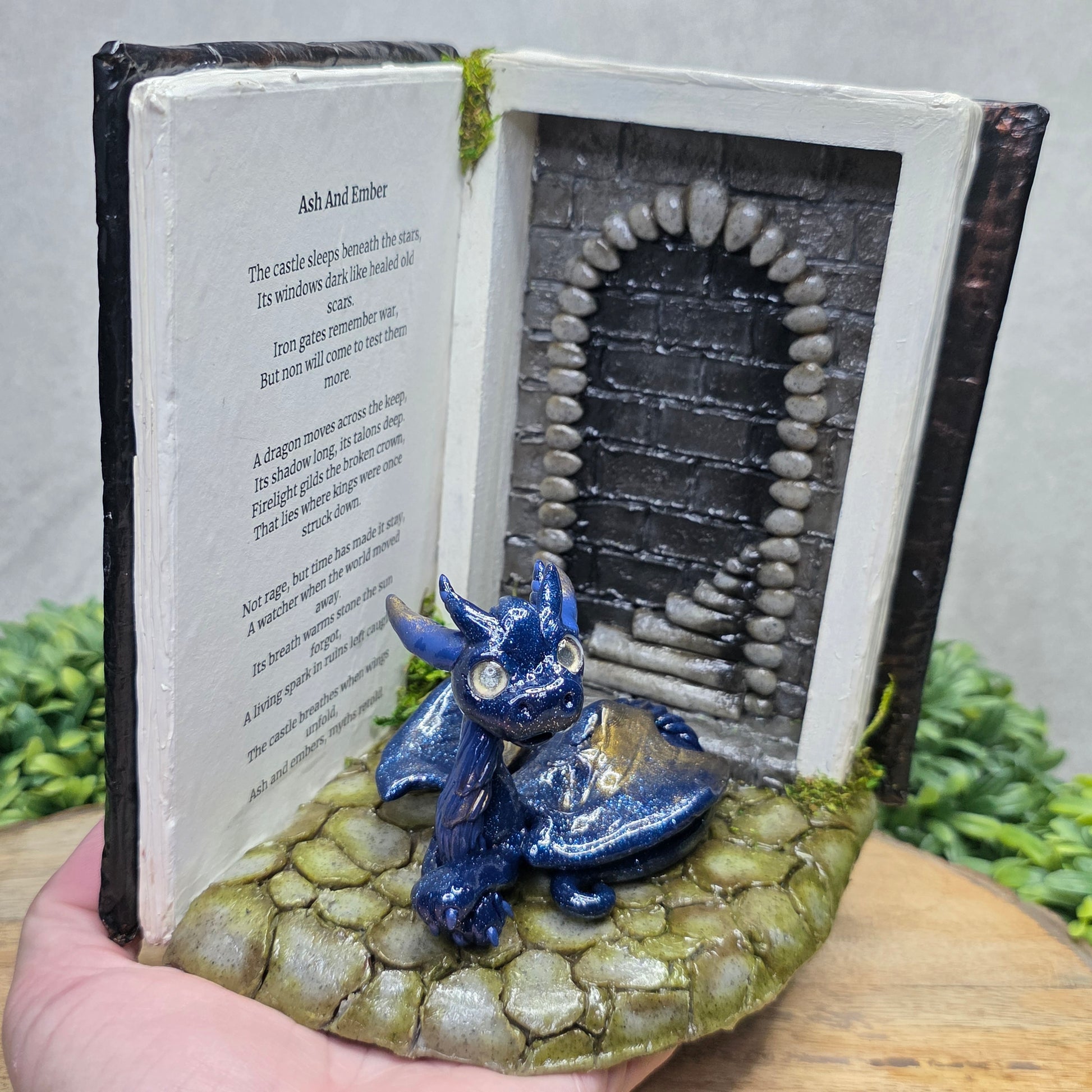 Hand holding a Book nook with dragon on a wood surface with greenery