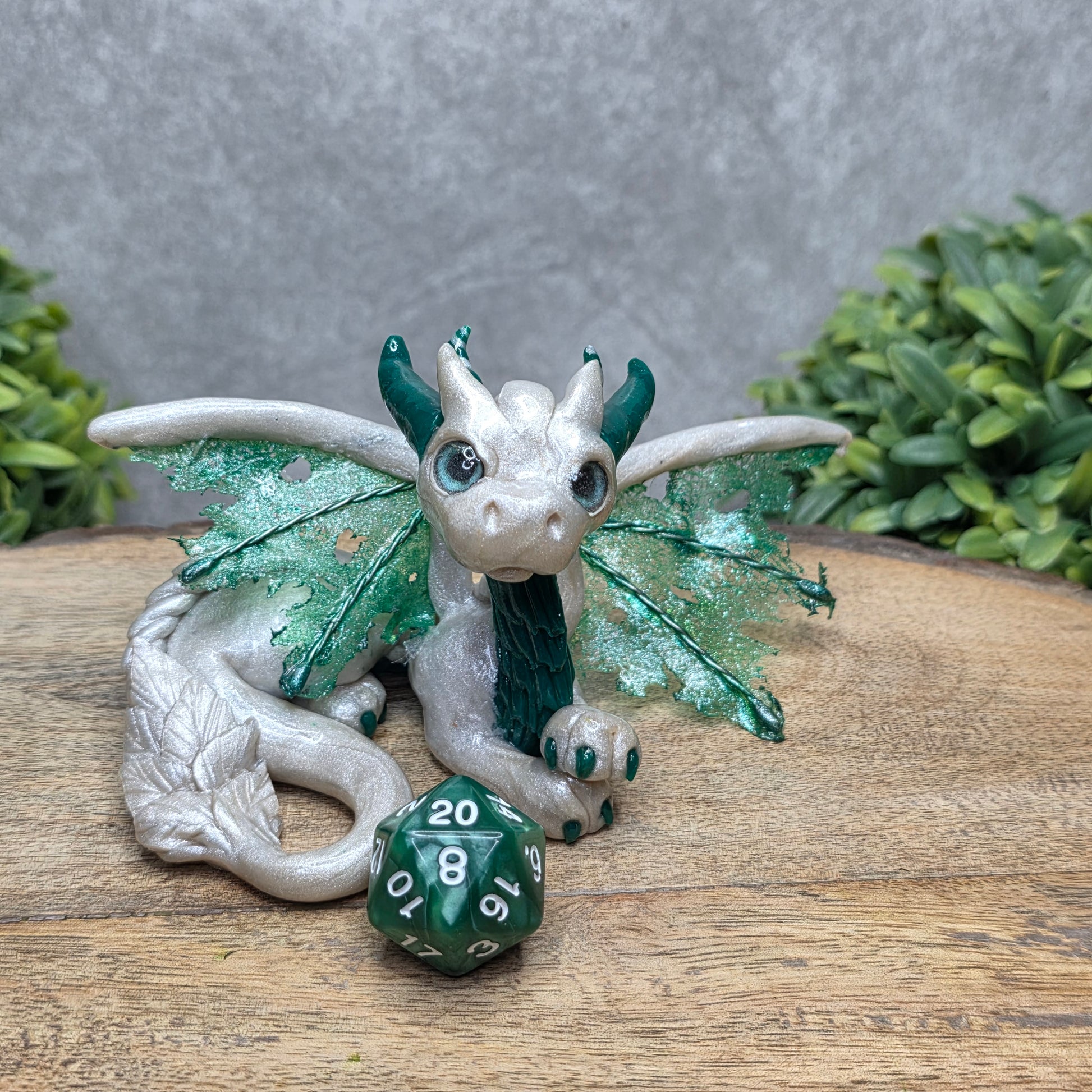 A clay dragon with a D20 dice against a wood surface with greenery.