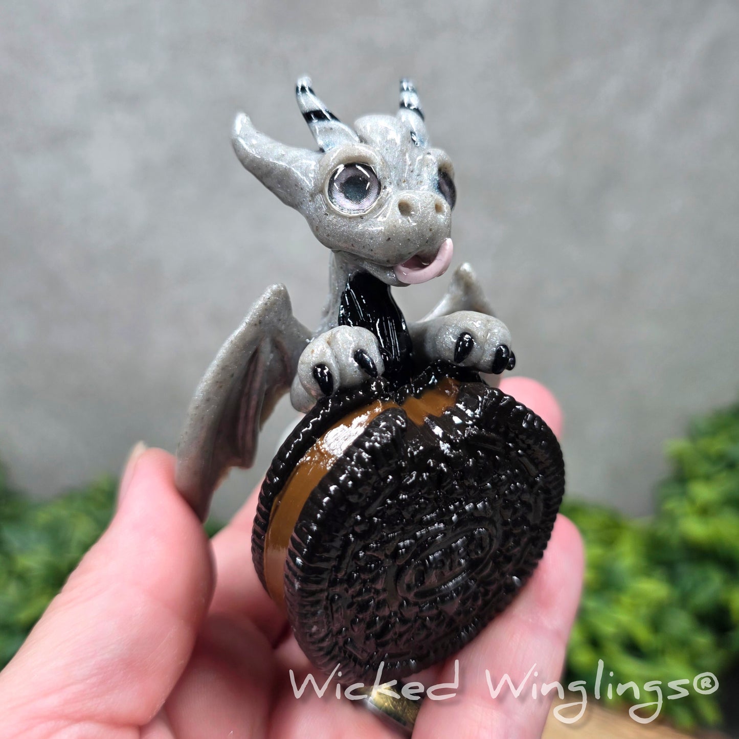 A hand holding a clay dragon with a cookie with a Wicked Winglings® watermark.