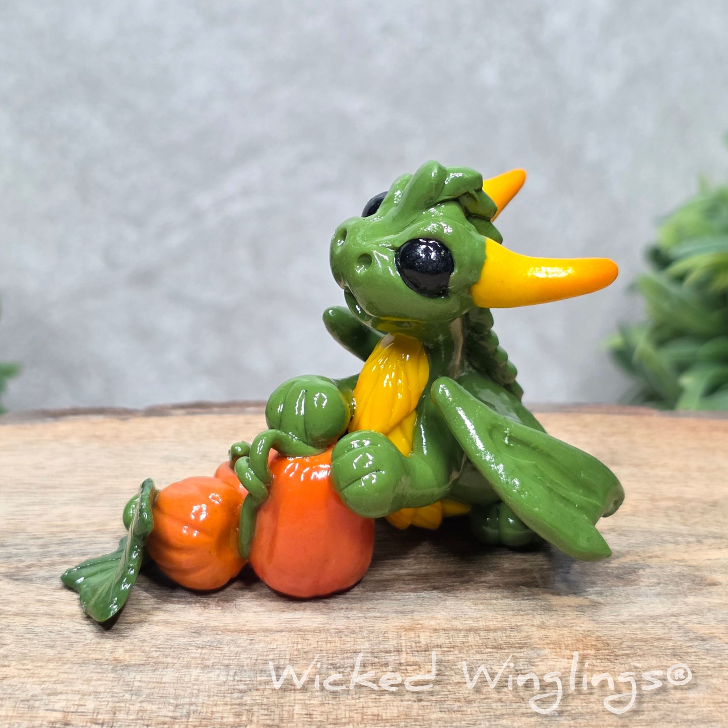 A baby clay dragon sculpture with pumpkins against a wood background with greenery and text Wicked Winglings