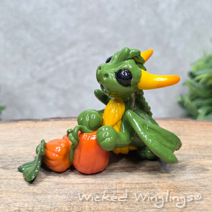 A baby clay dragon sculpture with pumpkins against a wood background with greenery and text Wicked Winglings