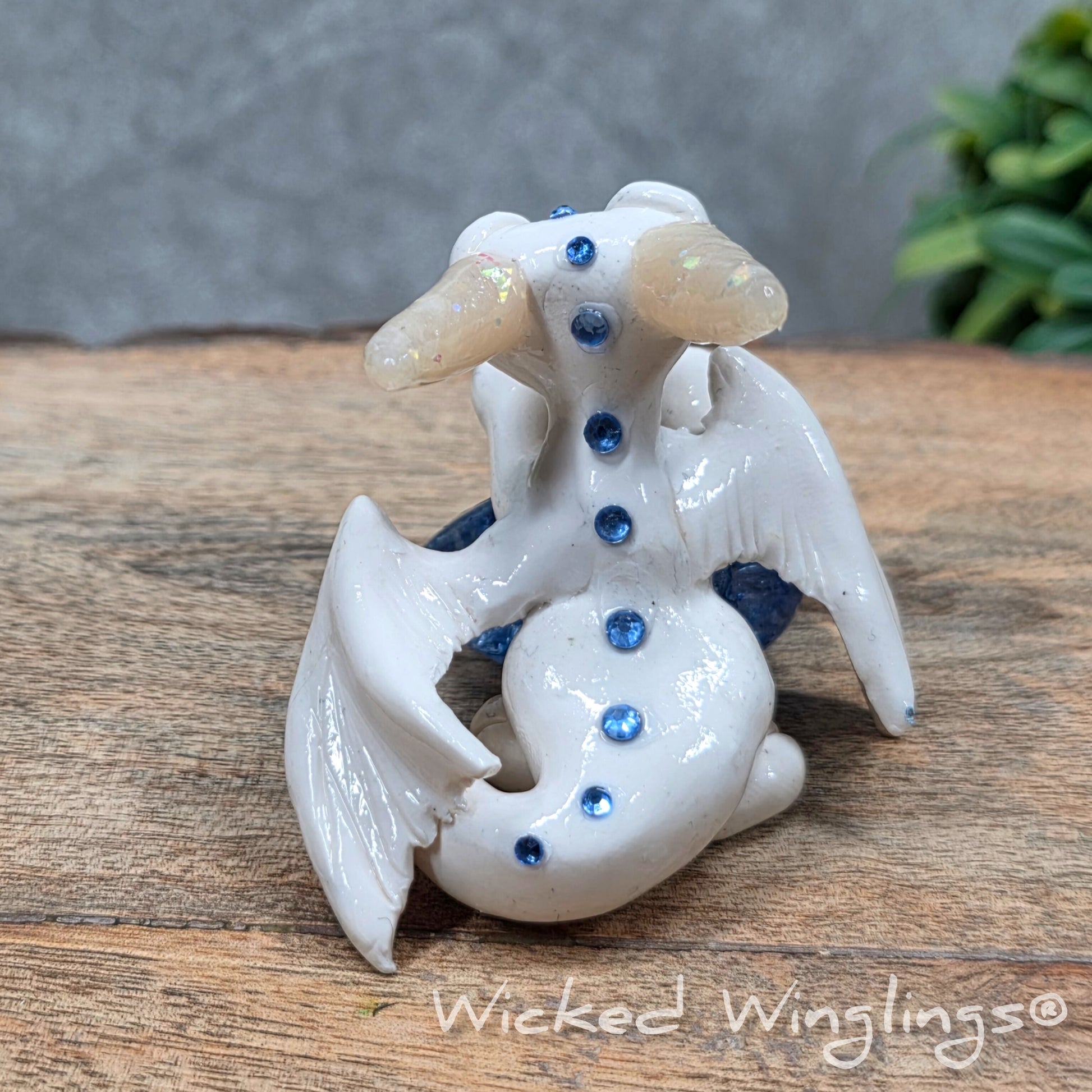 A baby clay dragon sculpture with a crackled quartz against a wood background with greenery and text Wicked Winglings