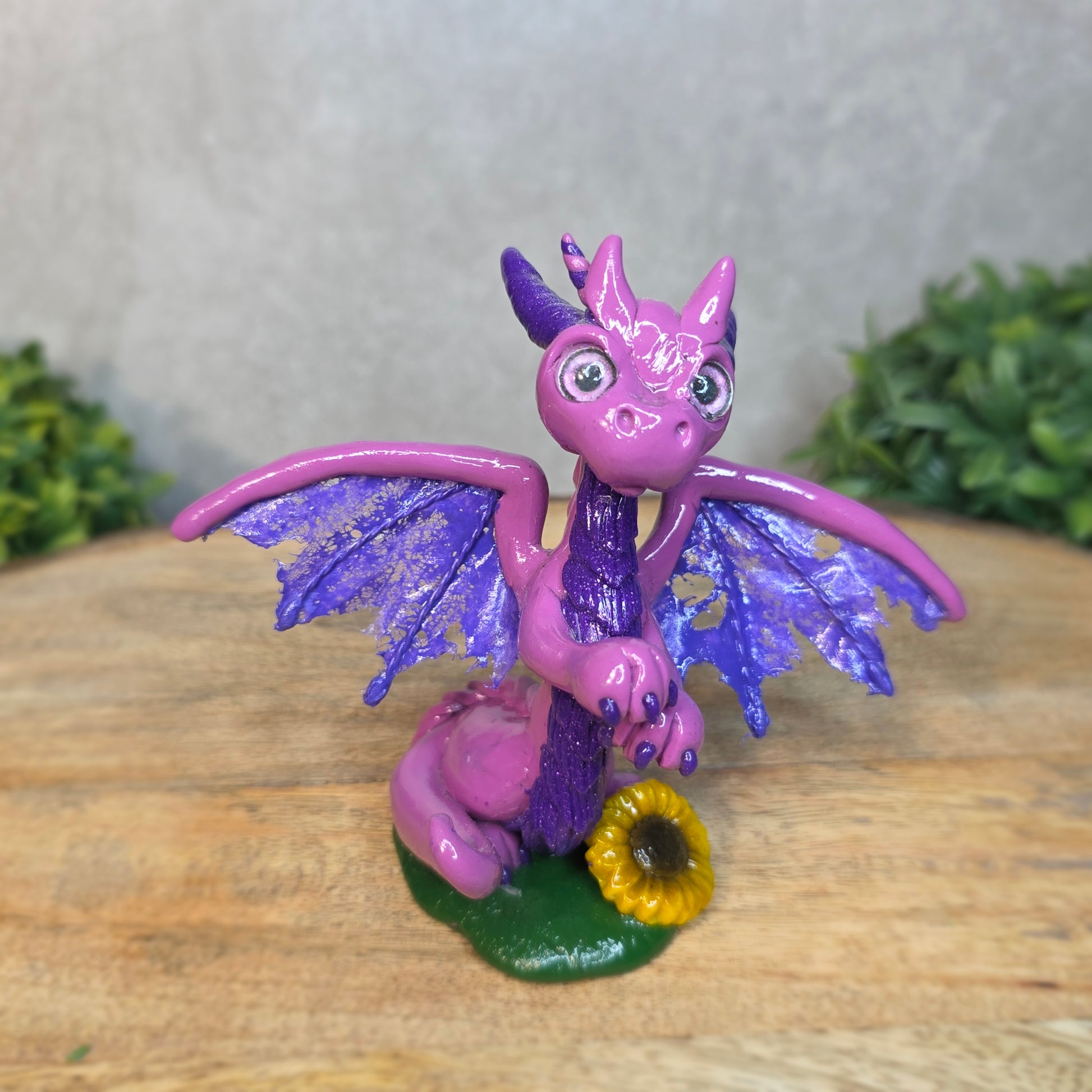 A clay dragon figurine with a sunflower on a wooden surface with a blurred green plant in the background