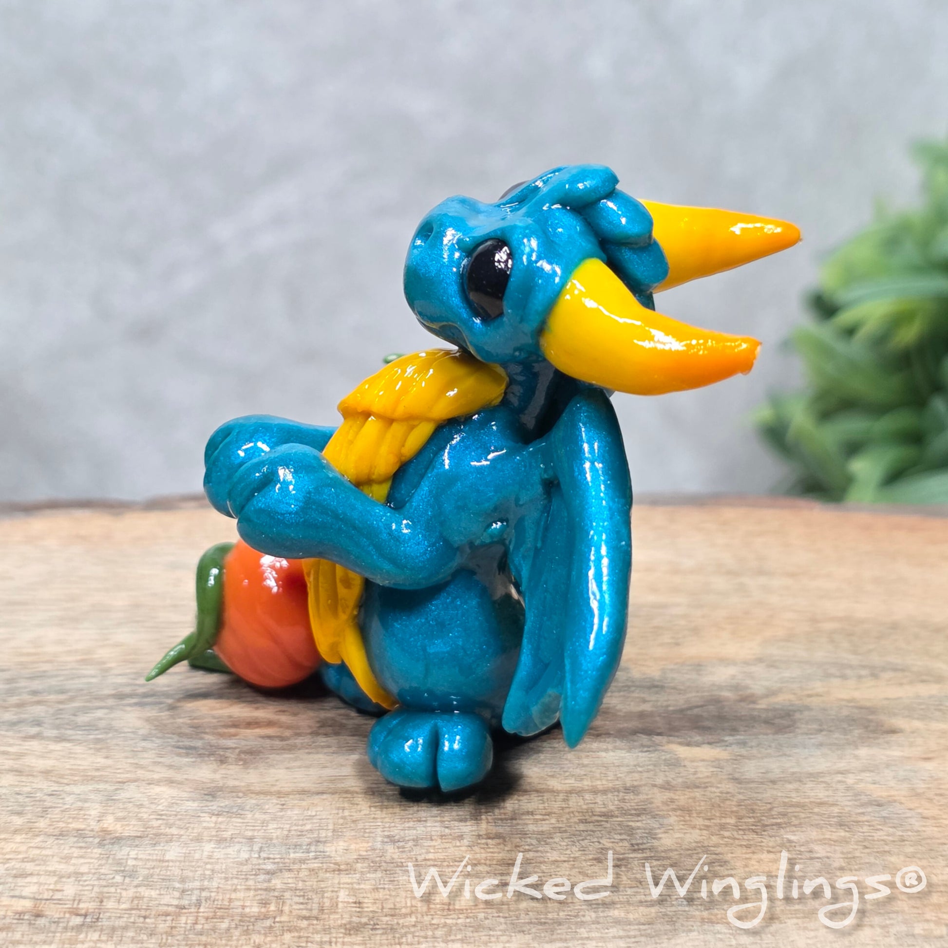 A baby clay dragon sculpture with pumpkins against a wood background with greenery and text Wicked Winglings