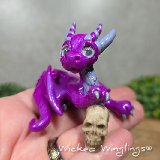 A hand holding a clay dragon with a skull against a blurred background with greenery. Text says Wicked Winglinng.
