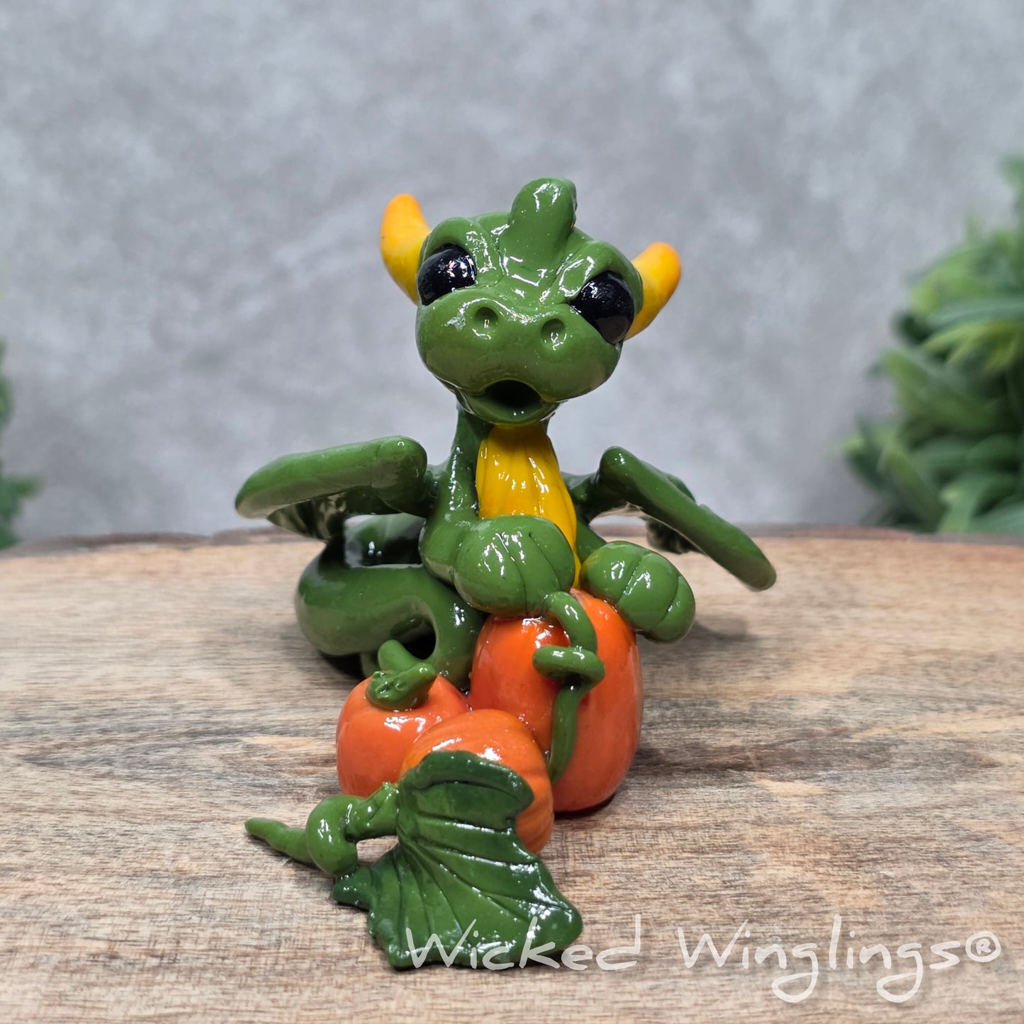 A baby clay dragon sculpture with pumpkins against a wood background with greenery and text Wicked Winglings