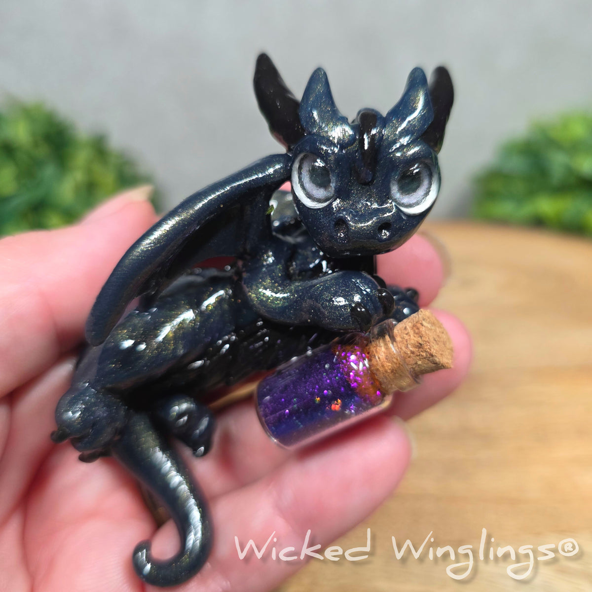 A hand holding a clay dragon with a potion bottle on a wood surface. Text reads Wicked Winglings.