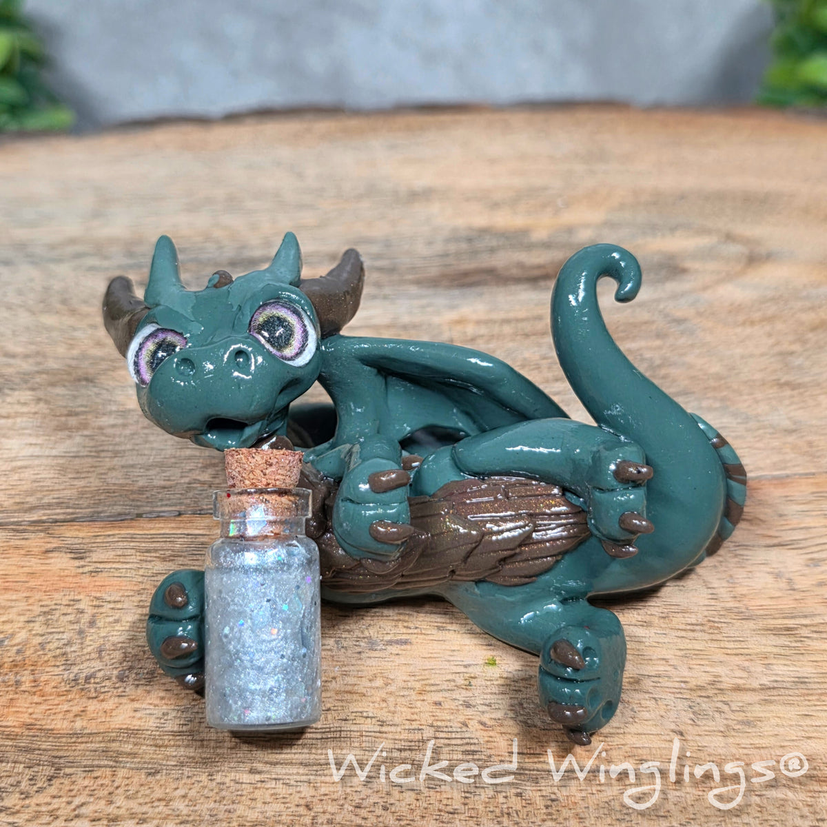 A clay dragon with a potion bottle on a wood surface. Text reads Wicked Winglings.