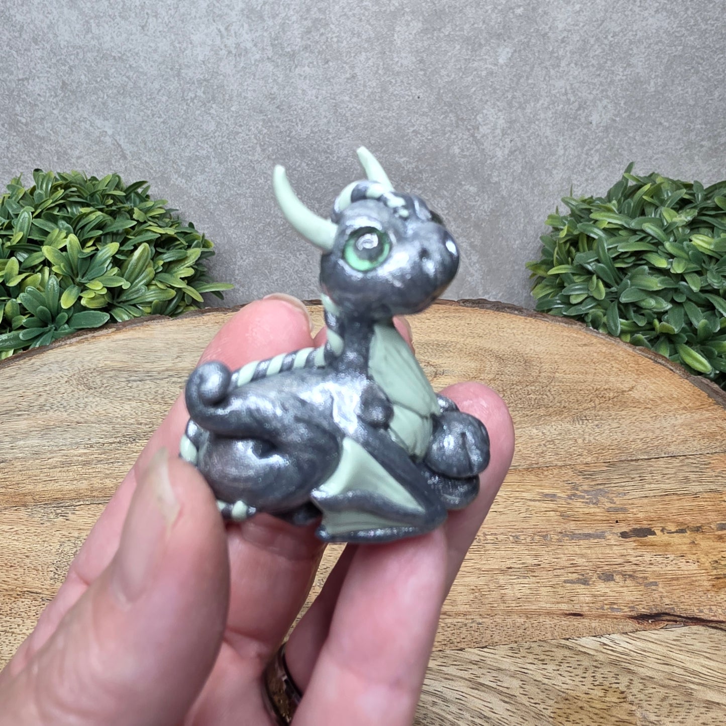 A hand holding a baby clay dragon against a wood background with greenery