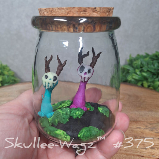 A glass jar with skull faced creatures and moss, held by a hand with a blurred green background. Text reads Skullee-Wagz 375