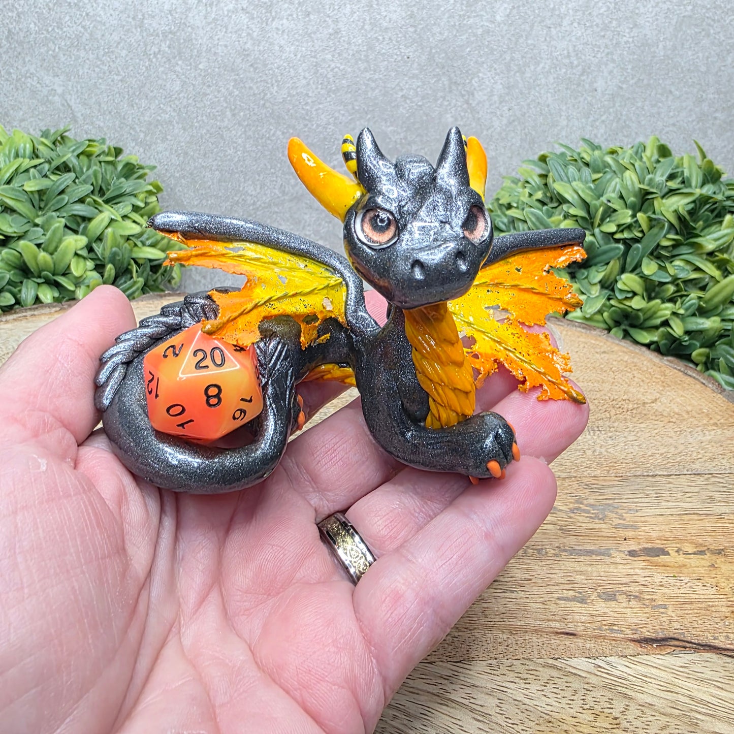Hand holding a clay dragon with a D20 dice against a wood surface with greenery.