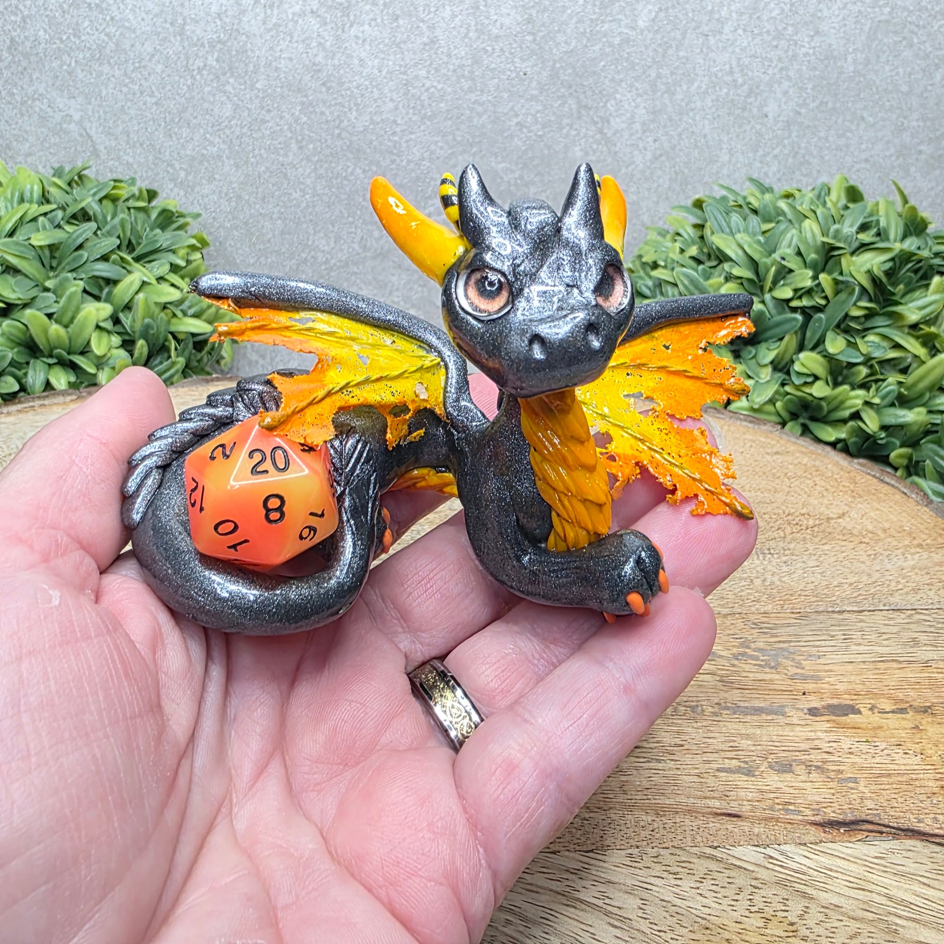 Hand holding a clay dragon with a D20 dice against a wood surface with greenery.