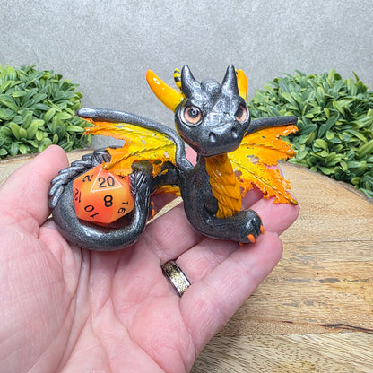 Hand holding a clay dragon with a D20 dice against a wood surface with greenery.