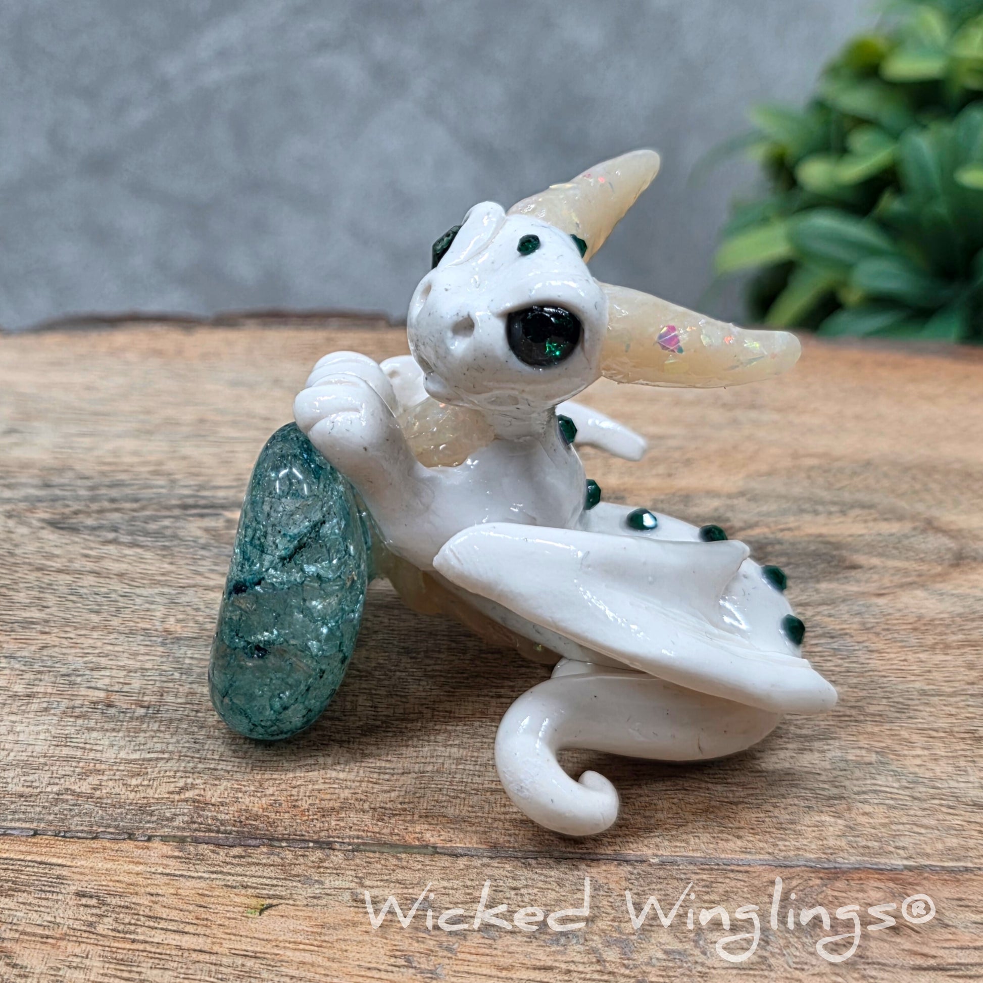 A baby clay dragon sculpture with a crackled quartz against a wood background with greenery and text Wicked Winglings
